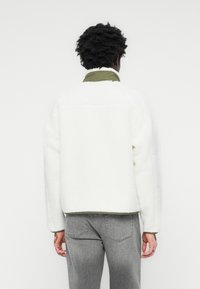 Hvit fleecejakke med grønn krage, med avslappet passform og teksturert stoff. Båret med grå jeans, som viser en uformell stil.
