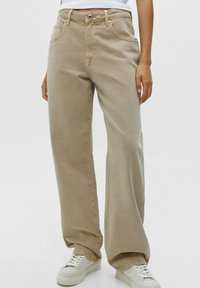 Jeans beige à jambes larges en coton avec une texture lisse, dotés d'une fermeture à bouton et zip, de poches latérales et de coutures minimales.