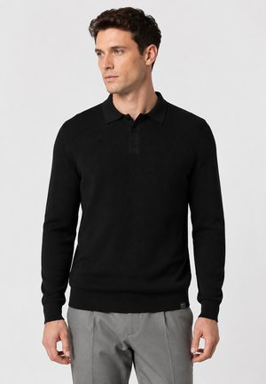 Mann trägt einen schwarzen langärmligen Polo-Pullover und graue Hose, steht und blickt leicht nach links vor einfachem Hintergrund.