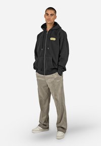 Juodas užtrauktukas megztinis pagamintas iš minkštos medžiagos, su priekinėmis kišenėmis ir kreminiu ženkleliu. Bežinės tekstūruotos kelnės ir baltos sportinės bateliai užbaigia aprangą.