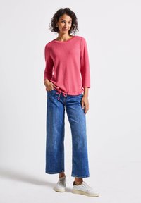 Korallrosa Stricktop mit Dreiviertelärmeln, verstellbarem Saum, kombiniert mit weit geschnittenen blauen Jeans und hellen Sneakern vor einem schlichten Hintergrund.