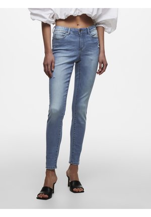 Vaqueros pitillo de color azul claro combinados con mules negros de tacón y punta abierta y una blusa corta blanca, mostrados desde el abdomen hasta los pies.