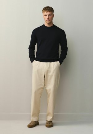 Tânăr stând pe lângă un perete simplu, purtând cămașă neagră cu mâneci lungi, pantaloni bej și pantofi maro, cu mâinile în buzunare.