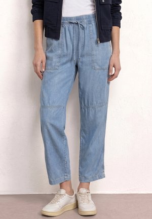 Person, die hellblaue Jeans-Jogginghosen mit elastischem Bund und weißen Sneakers trägt, stehend vor einer weiß strukturierten Wand.