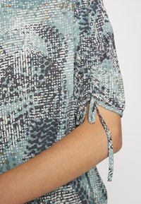 Blouse à manches courtes à motifs bleu et blanc avec cordon réglable noué à la manche au-dessus d'un bras plié.