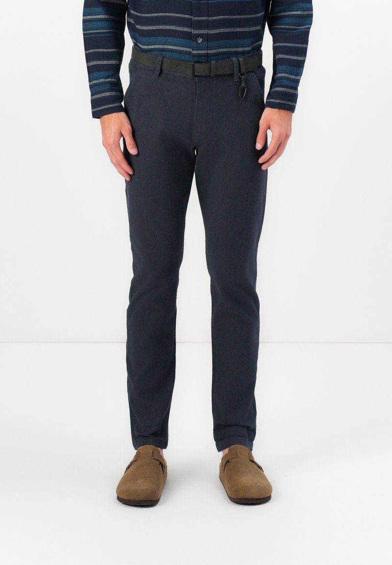 Tengeri kék pamut nadrág, slim fit szabással, fekete csattal ellátott övvel, barna velúrbőr slip-on cipőkkel párosítva.