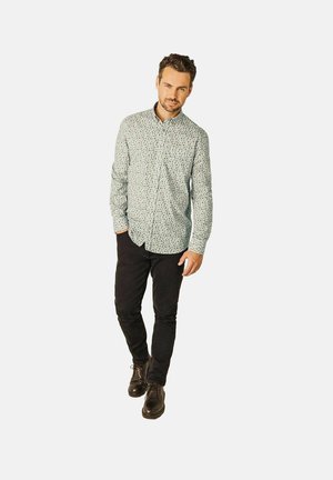 Man wearing a light gray patterned button-up shirt, black pants, and black lace-up shoes standing against a plain white background.