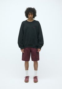 Sudadera negra con mangas raglán, pantalones cortos cargo burdeos con bolsillos laterales, calcetines blancos, y zapatos burdeos con suela texturizada.