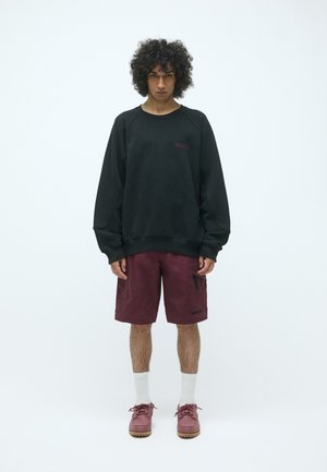Schwarzer Pullover mit Raglanärmeln, kastanienbraune Cargo-Shorts mit seitlichen Taschen, weiße Kniestrümpfe und kastanienbraune Schuhe mit strukturierter Sohle.