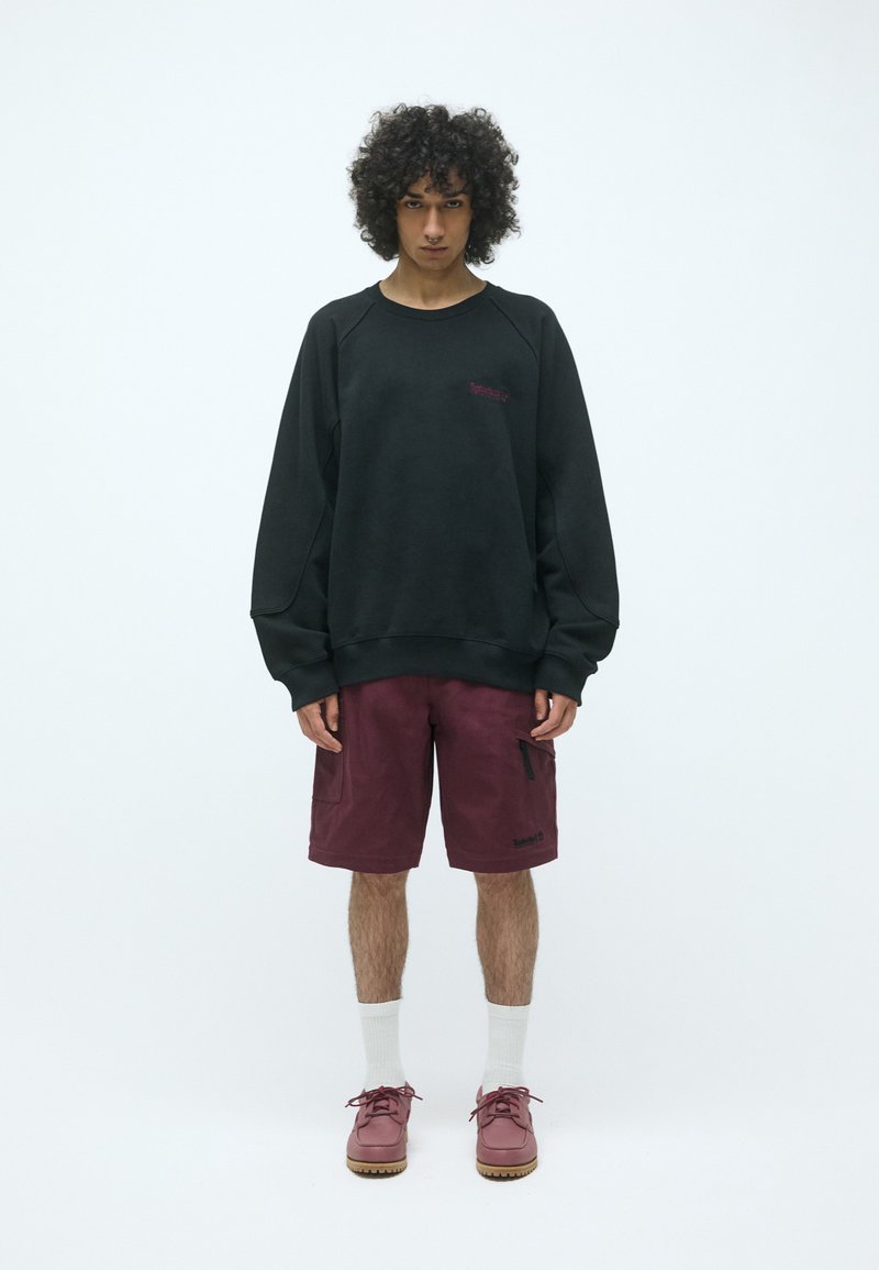 Sudadera negra con mangas raglán, pantalones cortos cargo burdeos con bolsillos laterales, calcetines blancos, y zapatos burdeos con suela texturizada.