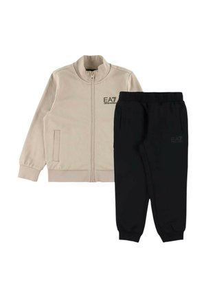 Giacca beige con zip e due tasche, pantaloni jogger con vita elastica nera, entrambi con loghi EA7 Emporio Armani discreti.
