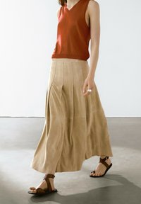 Gonna plissettata beige realizzata in tessuto morbido, abbinata a una canotta rossa senza maniche. Sandali marroni con fettucce minimal, che enfatizzano un look casual.