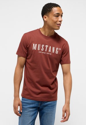 Bordó póló puha pamutból, kerek nyakkal és rövid ujjakkal. Elöl fehér szöveg található: "MUSTANG Jeans Company Since 1932".