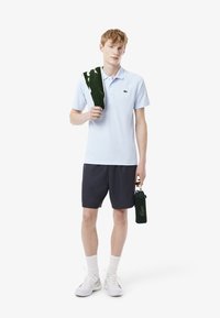 Ljusblå pikétröja med en liten logotyp, marinblå shorts, vita sneakers, hållande en grön handduk och en grön nätvattentank.