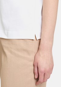 Haut en coton blanc avec un détail de fente sur le côté, associé à un tissu beige clair, montrant une texture lisse et des éléments de design simples.