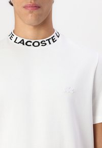 T-shirt blanc à manches courtes avec un col côtelé noir et blanc affichant "LACOSTE" et un petit logo de crocodile blanc sur la poitrine gauche.