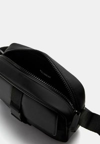 Borsa a tracolla nera in materiale sintetico con chiusura zip, una tracolla in tessuto e una texture liscia. Presenta una tasca frontale e un branding minimale.