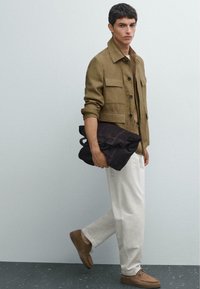 Bruine linnen knoopjasje met zakken, beige shirt, witte broek en bruine suède schoenen. Houdt een donkergrijze zachte stoffen tas vast.