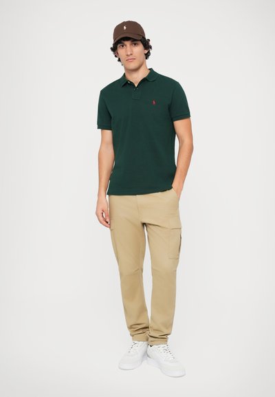Mörkgrön polo skjorta med en röd logotyp, beige cargobyxor med sidfickor och vita sneakers; modellen bär en brun keps.