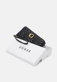 Schwarze gesteppte Geldbörse aus synthetischem Leder mit einem goldenen Logo und einem Handgelenkband, das in einer weißen Box liegt, die mit "GUESS" beschriftet ist.