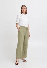 Chemise blanche à manches courtes avec des accents en dentelle, associée à un pantalon olive ample et des sandales noires à lanières.