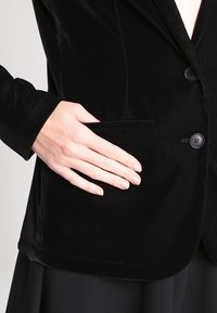 Černé sametové sako s jedním zapínacím knoflíkem, vykrojenými klopami a boční kapsou; hladká textura a střih na míru.