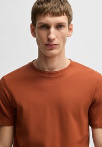 T-shirt a maniche corte, in tessuto texturizzato, di colore arancio ruggine, con scollatura tonda e finitura liscia. Design semplice senza decorazioni o motivi.