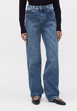 Modèle portant un jean bleu taille haute droit, un top noir à manches longues et des ballerines noires à bout carré sur fond blanc.