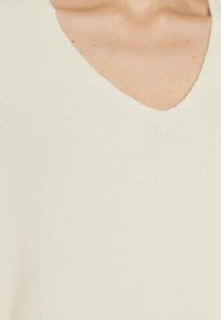 Gros plan d'un pull en maille blanc à col en V porté par une personne, montrant la texture et les détails du tissu.