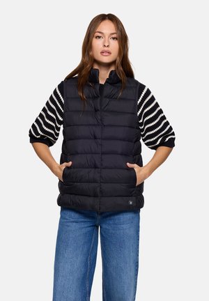 Chaleco negro acolchado con costuras horizontales y bolsillos laterales. Lleva puesto un suéter de punto con rayas en blanco y negro, que tiene un cuello alto y cremallera.