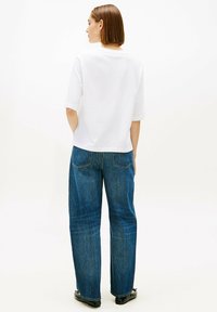 Camisa blanca de manga corta combinada con vaqueros azules sueltos de mezclilla, con corte recto y bolsillos parche en la parte trasera, vista desde atrás.