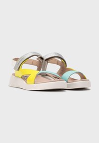 Sandalias coloridas con una correa metálica plateada, acentos en amarillo y azul verdoso, diseño de punta abierta y una suela blanca acolchada.