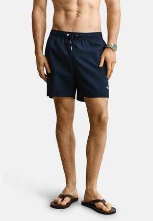 Homme portant un short de bain bleu marine et des tongs noires, debout avec les mains détendues, montre au poignet gauche, torse nu visible.
