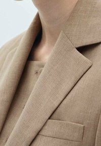 Blazer en lin beige à revers cranté porté sur un haut beige assorti, montrant le col et la partie supérieure de la poitrine.
