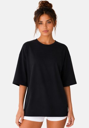 Jonge vrouw met bruin haar in een knot, draagt een los zwart t-shirt en witte shorts, staand tegen een effen witte achtergrond.