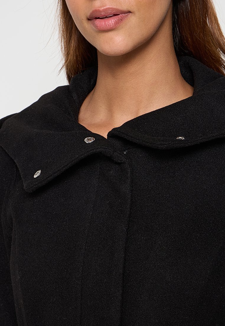 Manteau en laine noire avec un grand col, doté de boutons-pression et d'une texture douce. Gros plan sur le détail du col et de l'encolure.