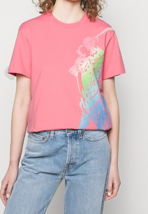 Personne portant un t-shirt rose avec un motif abstrait multicolore sur le côté, associé à un jean taille haute bleu clair.