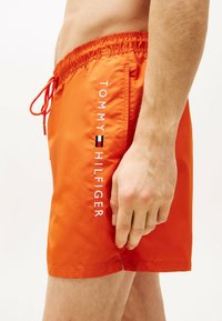 Pantaloni da bagno arancioni realizzati in tessuto leggero, con un elastico in vita e un logo bianco a contrasto con accenti blu e rossi.