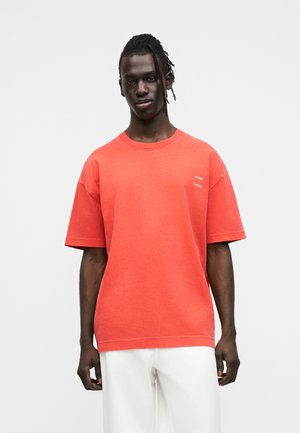 T-shirt rouge oversize en coton. Manches courtes, encolure ronde, avec un petit texte imprimé sur la poitrine gauche. Texture lisse.