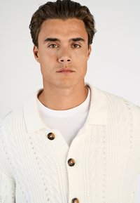 Jonge man met bruine ogen en donker haar draagt een wit, gestructureerd vest met schildpadknopen over een wit T-shirt met ronde hals.