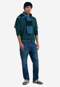 Lässiges Outfit mit grünem Kapuzenpullover, blauem Schal und blauer Jeans. Die Schuhe sind grau mit weißen Akzenten. Schlichtes Design und bequeme Passform.
