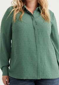 Grön skjorta med knappar, mönstrad med en textur, långa ärmar och krage. Tyget verkar vara lättviktigt med små upphöjda prickar överallt.