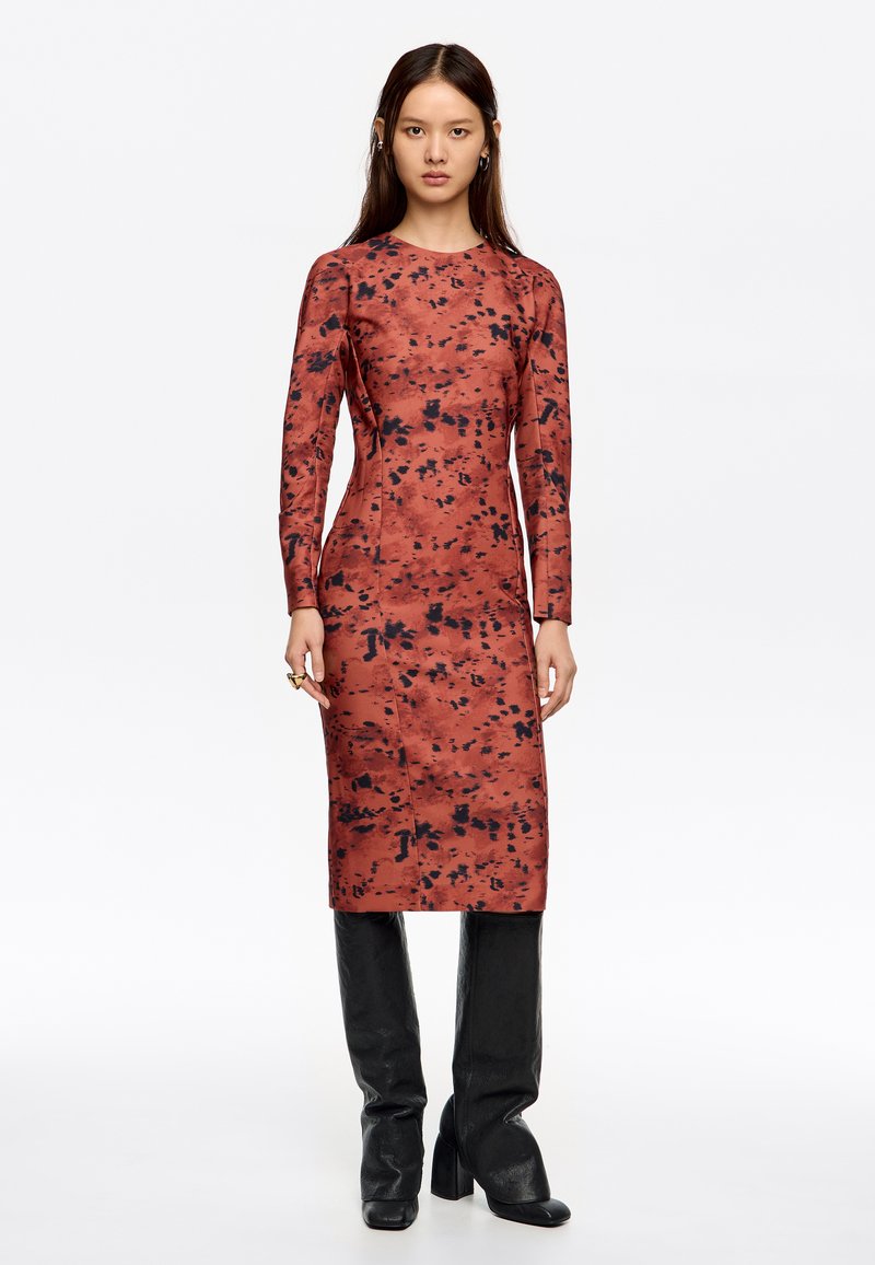 Vestido de manga larga con un patrón en rojo y negro, de diseño ajustado, hasta la rodilla, hecho de un material suave. Llevado con botas de tacón negro.