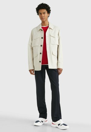 Chaqueta beige clara con cuatro bolsillos delanteros y botones negros, usada sobre una camisa roja, combinada con pantalones azul marino y zapatillas blancas.
