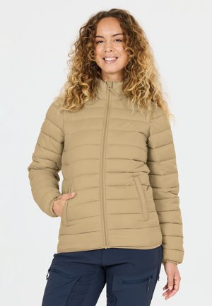 Glimlachende vrouw met krullend haar, gekleed in een beige doorgestikte jas en marineblauwe cargobroek, staand met één hand in haar zak.