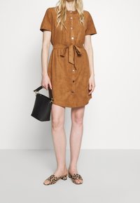 Robe chemise en suède marron avec des manches courtes, des boutons à l'avant et une taille cintrée. Associée à des mules à motif léopard et un sac seau noir.