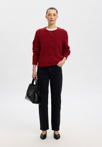 Raudonas megztas kardiganas su apvalia iškirpte ir juodais mygtukais, derinamas su juodais tiesiais džinsais ir juoda rankine. Lygūs odiniai batai.