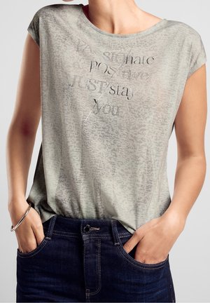 Kvinde iført lyse mønstrede kortærmede skjorte med teksten "Passionate & Positive Just stay you", hænder i lommerne på mørkeblå jeans, sølvarmbånd.