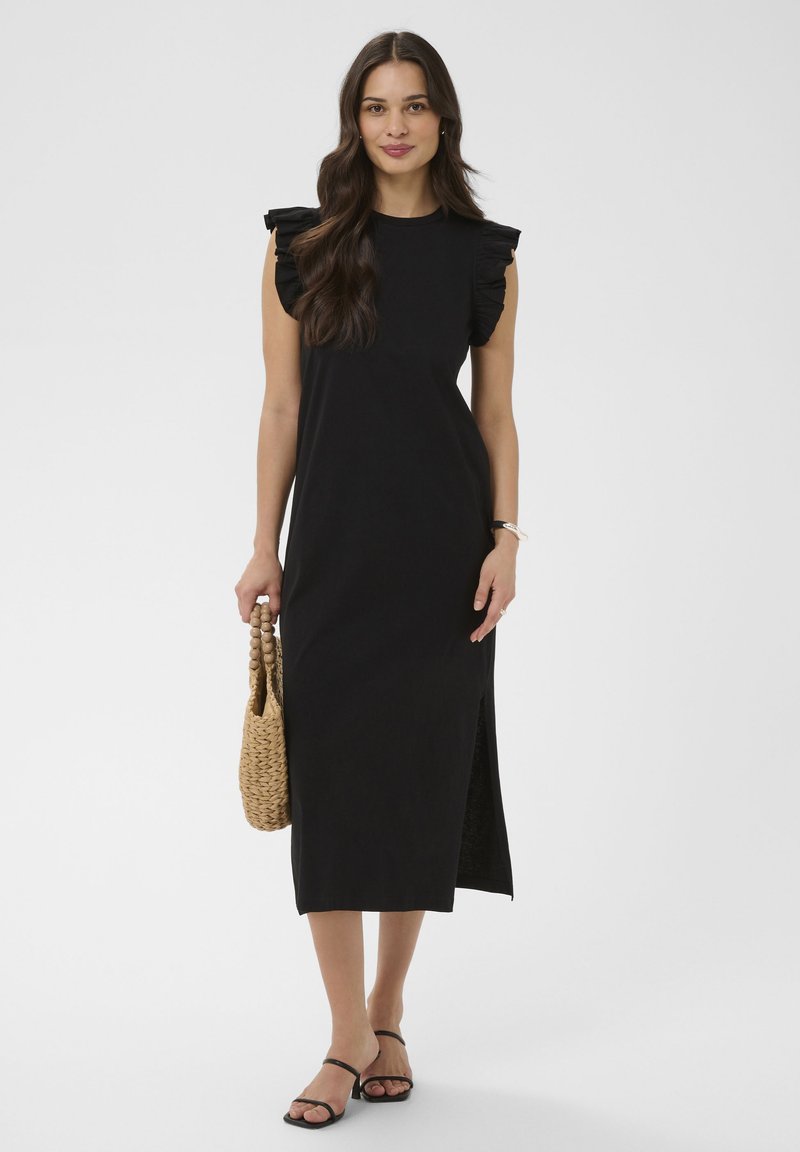 Femme portant une robe midi noire sans manches avec des épaules à volants, tenant un sac tissé et chaussée de sandales noires à fines lanières.