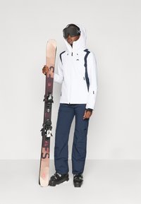 Balta slidinėjimo striukė su tamsiai mėlynomis detalėmis, tamsiai mėlynos slidinėjimo kelnės, juodi slidinėjimo batai ir slidės su laikikliais rankoje. Modelis dėvi slidinėjimo akinius.